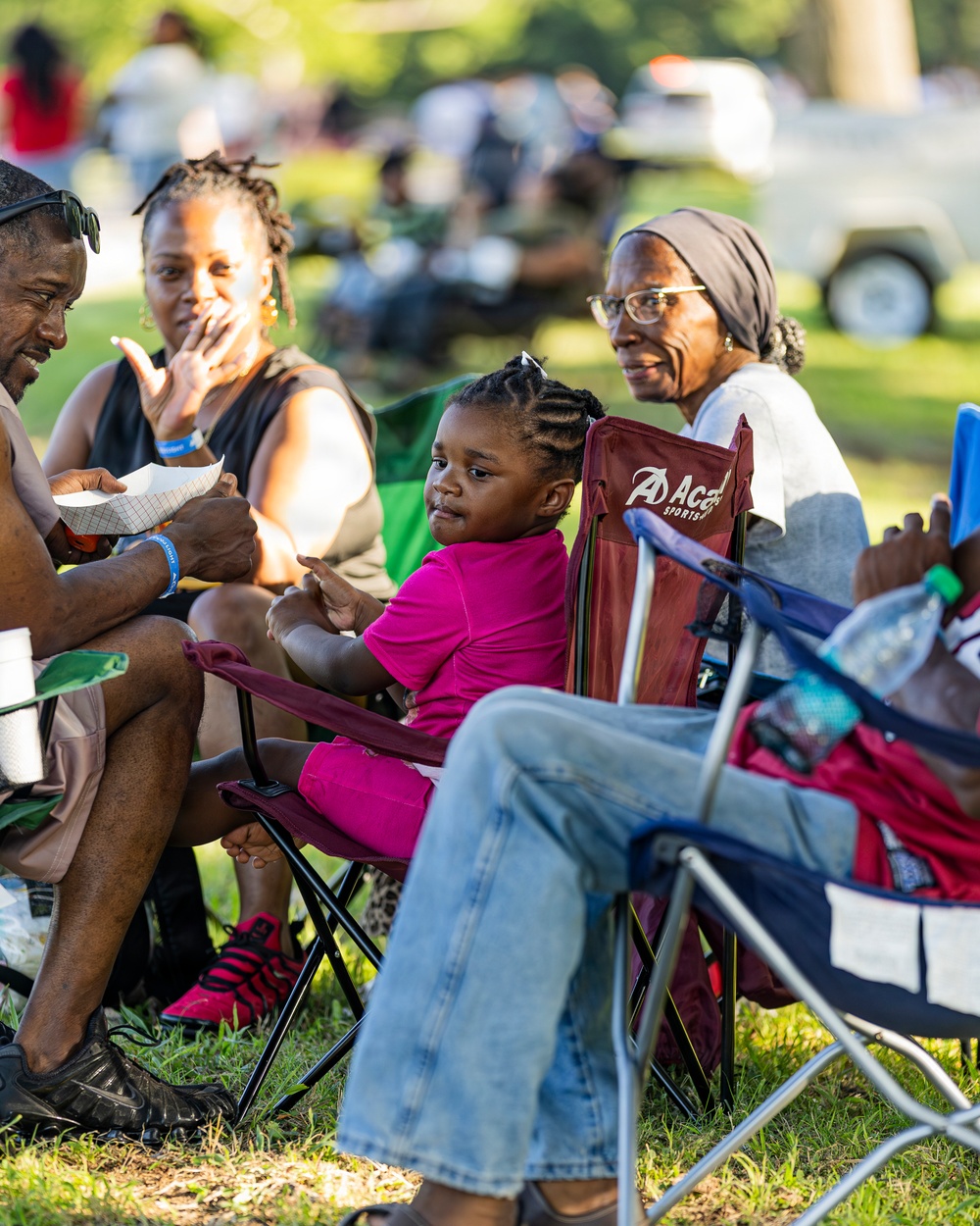 A group of people, appearing to be a multi-generational family, are seated in portable folding chairs on grass at an outdoor event. The group includes an older woman wearing glasses and a head wrap, a younger adult woman, a man, and a young child dressed in pink. The background shows a blurred crowd and parked vehicles, suggesting a community gathering or outdoor festival.