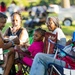 A group of people, appearing to be a multi-generational family, are seated in portable folding chairs on grass at an outdoor event. The group includes an older woman wearing glasses and a head wrap, a younger adult woman, a man, and a young child dressed in pink. The background shows a blurred crowd and parked vehicles, suggesting a community gathering or outdoor festival.