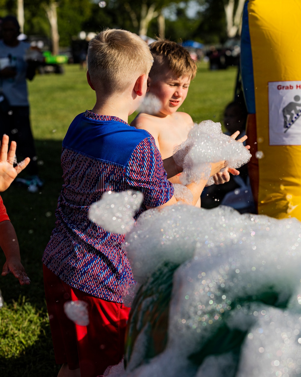 Two young boys are playing at an outdoor foam/bubble machine attraction at what appears to be a community or carnival event. One boy is wearing a blue patterned shirt and red shorts, while the other is shirtless, and both are interacting with large amounts of white foam. The setting is a grassy park area with trees and other event activities visible in the background.
