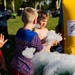 Two young boys are playing at an outdoor foam/bubble machine attraction at what appears to be a community or carnival event. One boy is wearing a blue patterned shirt and red shorts, while the other is shirtless, and both are interacting with large amounts of white foam. The setting is a grassy park area with trees and other event activities visible in the background.
