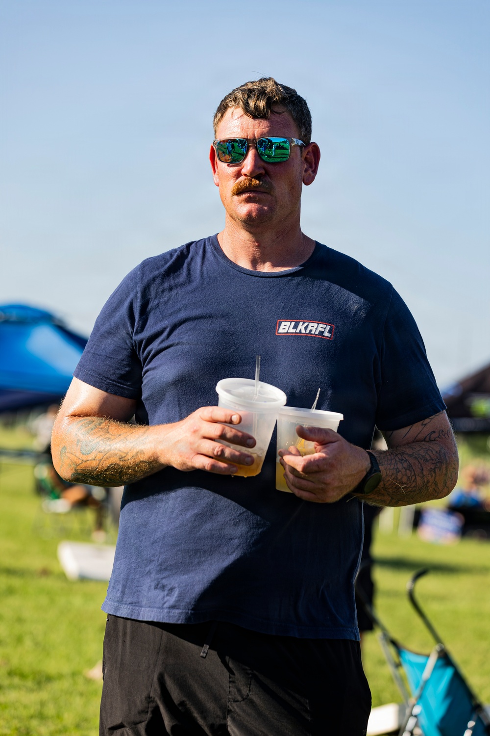 A man with a mustache and green-mirrored sunglasses stands outdoors at what appears to be a casual outdoor event or festival. He is holding two plastic cups with straws containing a light-colored beverage. He is wearing a navy blue t-shirt branded 'BLKAFL' and has visible tattoos on both forearms.