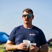 A man with a mustache and green-mirrored sunglasses stands outdoors at what appears to be a casual outdoor event or festival. He is holding two plastic cups with straws containing a light-colored beverage. He is wearing a navy blue t-shirt branded 'BLKAFL' and has visible tattoos on both forearms.