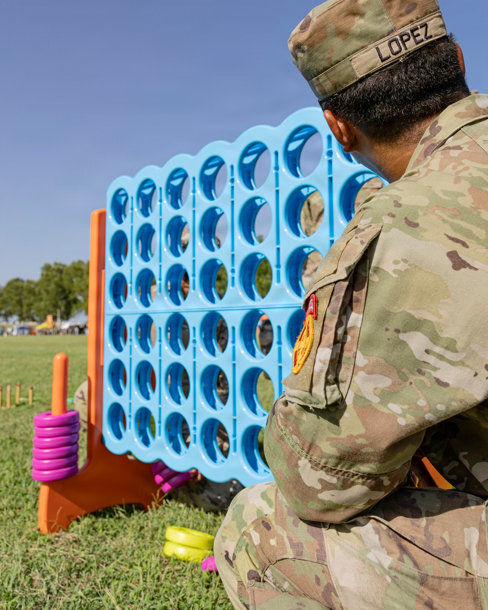 A U.S. Army soldier named Lopez, wearing OCP (Operational Camouflage Pattern) uniform, crouches on a grass field examining a large oversized Connect Four game board. The blue plastic game grid is mounted on an orange stand, with colorful rings (pink, yellow) visible nearby as game pieces. The setting appears to be an outdoor recreational or community event on a military installation.