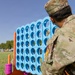 A U.S. Army soldier named Lopez, wearing OCP (Operational Camouflage Pattern) uniform, crouches on a grass field examining a large oversized Connect Four game board. The blue plastic game grid is mounted on an orange stand, with colorful rings (pink, yellow) visible nearby as game pieces. The setting appears to be an outdoor recreational or community event on a military installation.