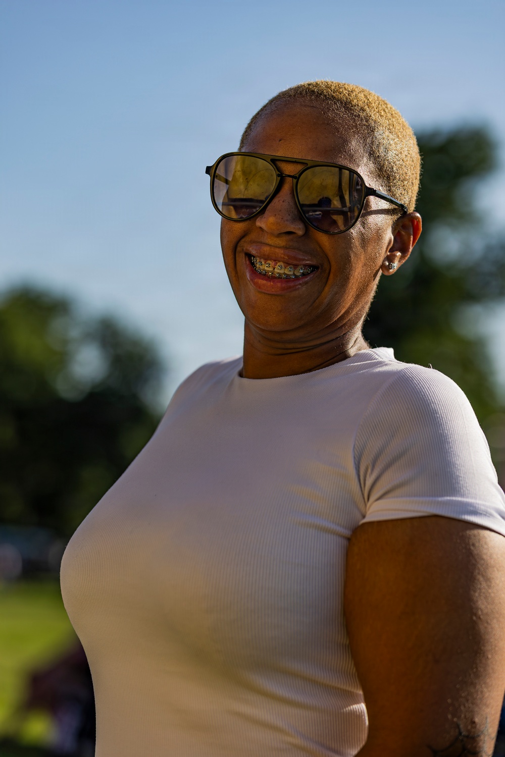 A woman is photographed outdoors in natural sunlight, smiling broadly. She has short blonde-dyed hair, wears aviator-style sunglasses with amber lenses, dental braces, and a white ribbed short-sleeve t-shirt. The background shows a blurred outdoor setting with green trees and blue sky.