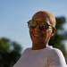 A woman is photographed outdoors in natural sunlight, smiling broadly. She has short blonde-dyed hair, wears aviator-style sunglasses with amber lenses, dental braces, and a white ribbed short-sleeve t-shirt. The background shows a blurred outdoor setting with green trees and blue sky.