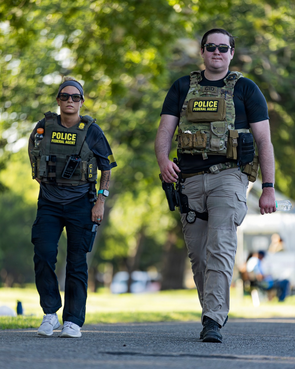 Two federal law enforcement agents wearing tactical gear are walking along a paved path in what appears to be a park or outdoor public area. Both agents are wearing plate carriers labeled 'POLICE FEDERAL AGENT' with 'CID' markings, sunglasses, and are visibly armed. The image is sharp and well-lit with a blurred green tree canopy in the background.