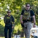 Two federal law enforcement agents wearing tactical gear are walking along a paved path in what appears to be a park or outdoor public area. Both agents are wearing plate carriers labeled 'POLICE FEDERAL AGENT' with 'CID' markings, sunglasses, and are visibly armed. The image is sharp and well-lit with a blurred green tree canopy in the background.
