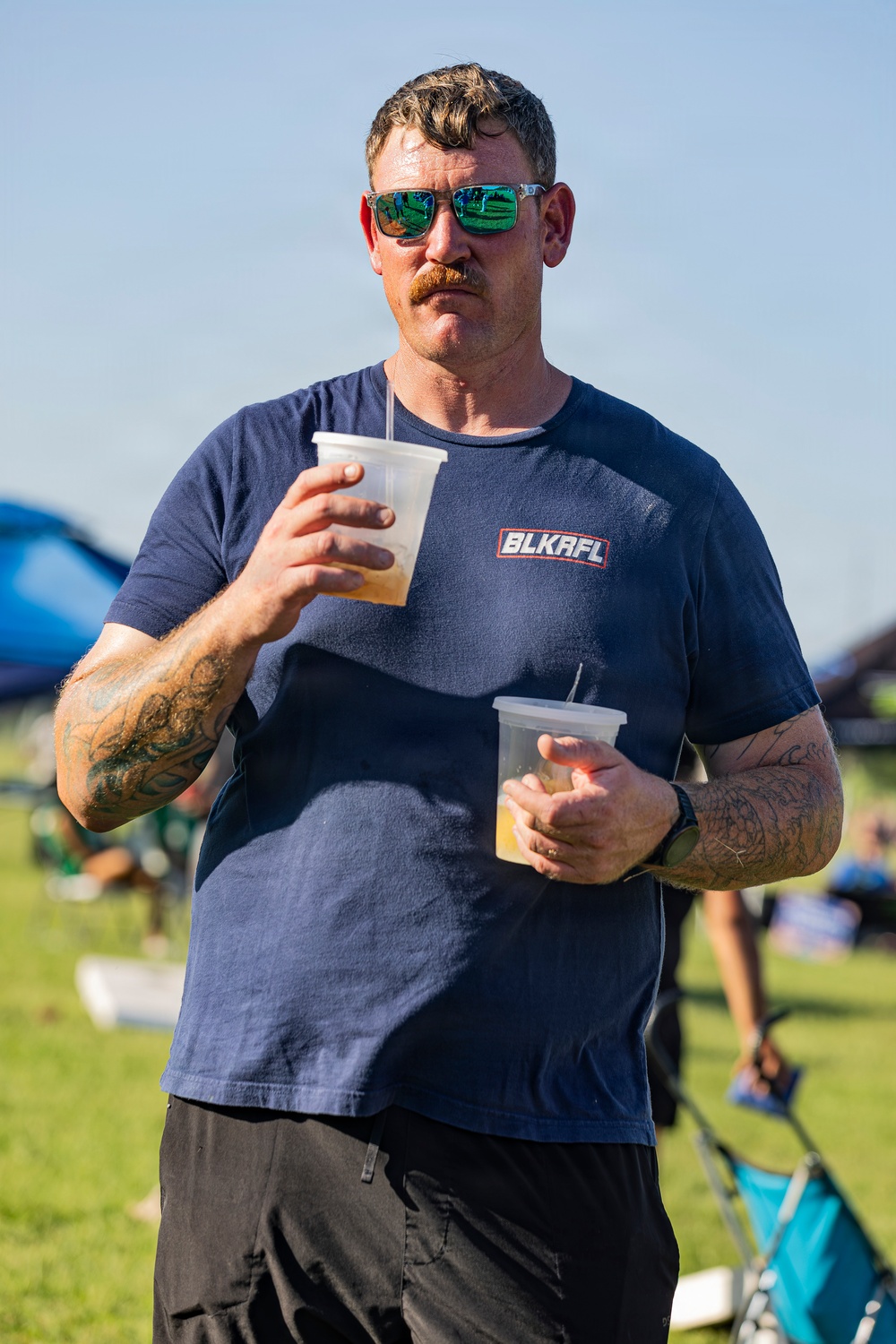 A middle-aged man with a mustache and green mirrored sunglasses stands outdoors at what appears to be a casual outdoor event or festival. He is carrying two plastic cups with lids, likely containing beverages. He wears a navy blue t-shirt branded 'BLKAFL' and has visible tattoos on both arms.