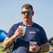 A middle-aged man with a mustache and green mirrored sunglasses stands outdoors at what appears to be a casual outdoor event or festival. He is carrying two plastic cups with lids, likely containing beverages. He wears a navy blue t-shirt branded 'BLKAFL' and has visible tattoos on both arms.