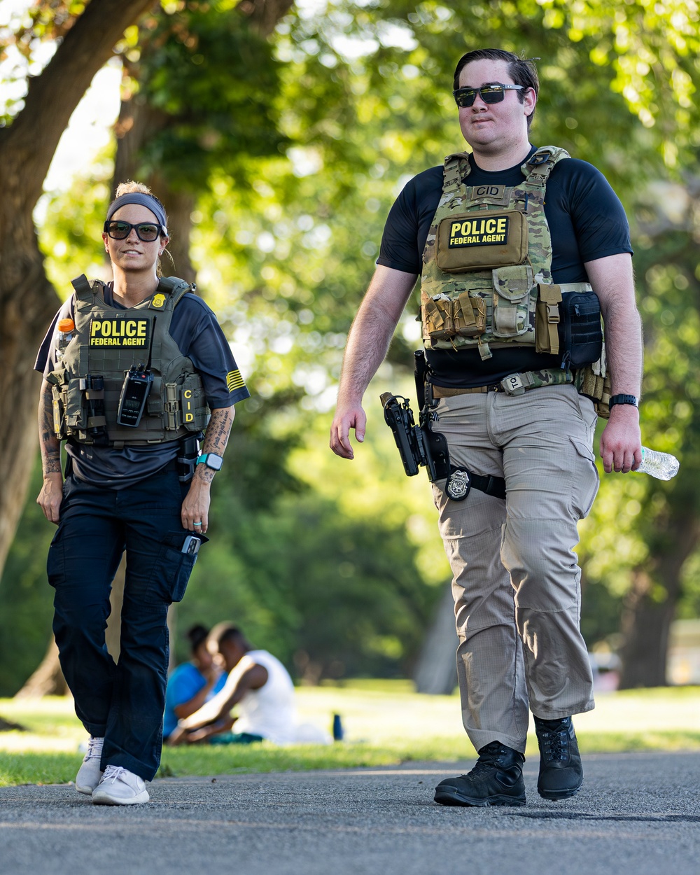 Two federal law enforcement agents wearing tactical gear are walking along a paved path in what appears to be a public park. Both wear plate carriers labeled 'POLICE FEDERAL AGENT' with 'CID' markings, and are visibly armed. The setting is outdoors with trees and civilians visible in the background.