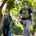 Two federal law enforcement agents wearing tactical gear are walking along a paved path in what appears to be a public park. Both wear plate carriers labeled 'POLICE FEDERAL AGENT' with 'CID' markings, and are visibly armed. The setting is outdoors with trees and civilians visible in the background.