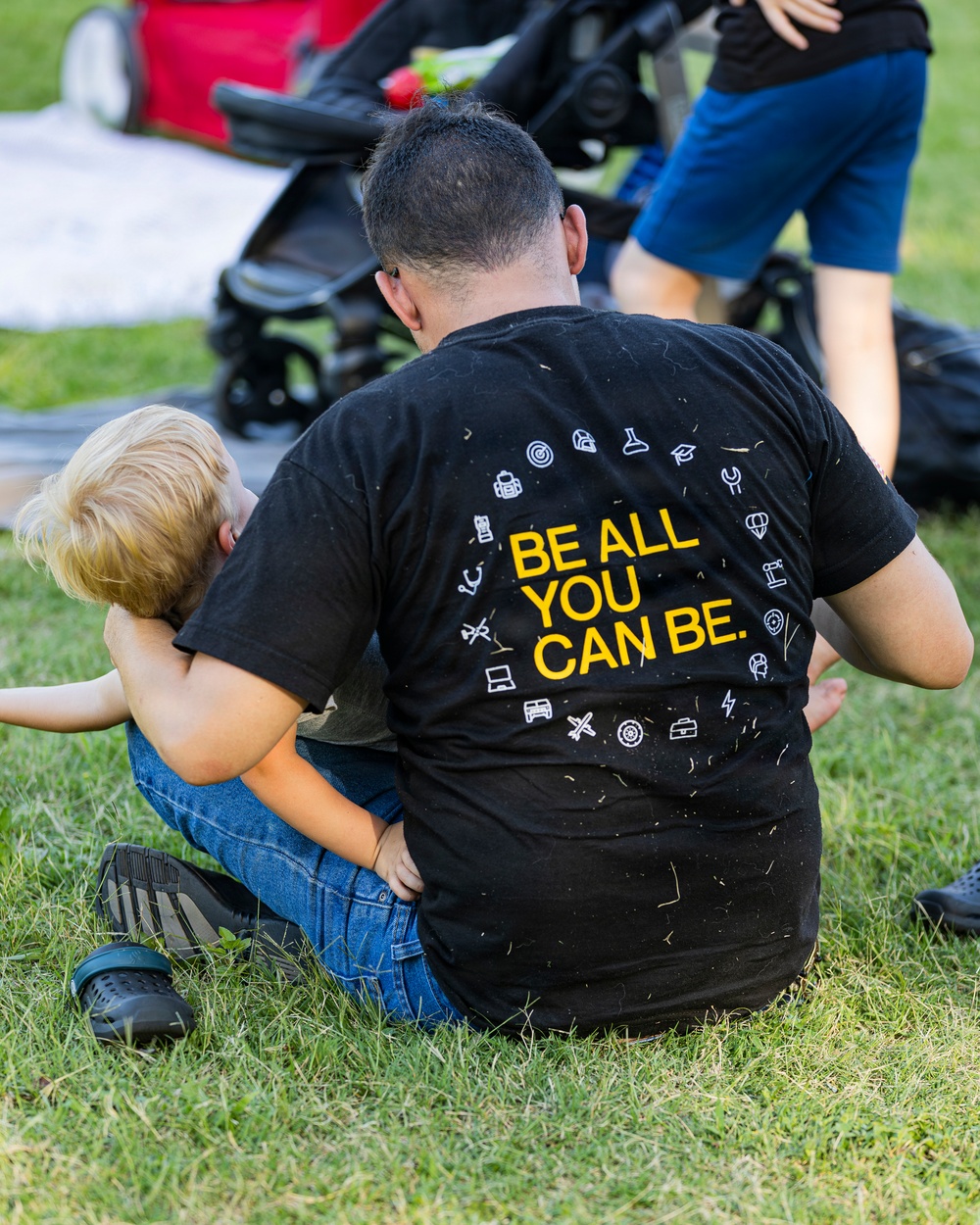 An adult sits on grass wearing a black t-shirt with 'BE ALL YOU CAN BE.' printed in yellow on the back, surrounded by various white icons. A young blonde child leans against the adult's side. The setting appears to be an outdoor public gathering or event, with a stroller and other people visible in the background.