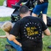 An adult sits on grass wearing a black t-shirt with 'BE ALL YOU CAN BE.' printed in yellow on the back, surrounded by various white icons. A young blonde child leans against the adult's side. The setting appears to be an outdoor public gathering or event, with a stroller and other people visible in the background.