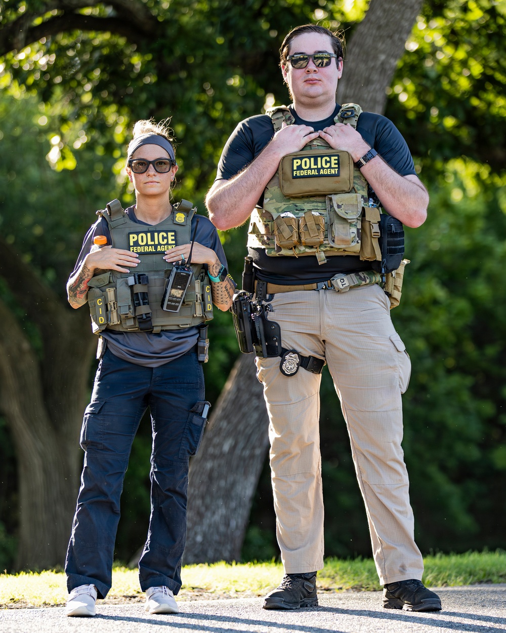 Two individuals wearing tactical gear and body armor labeled 'POLICE FEDERAL AGENT' are standing outdoors in a wooded area. Both wear sunglasses and are equipped with holsters, radios, badges, and MOLLE-style plate carriers. The image appears to be a professional or posed photograph taken in daylight.