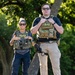 Two individuals wearing tactical gear and body armor labeled 'POLICE FEDERAL AGENT' are standing outdoors in a wooded area. Both wear sunglasses and are equipped with holsters, radios, badges, and MOLLE-style plate carriers. The image appears to be a professional or posed photograph taken in daylight.