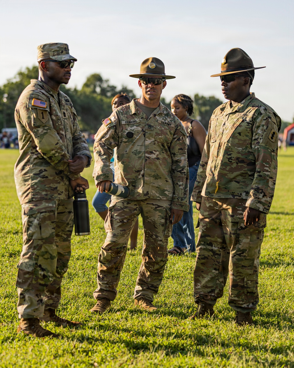 Three U.S. Army soldiers in Operational Camouflage Pattern (OCP) uniforms are standing on a grass field engaged in conversation. Two of the soldiers are wearing Drill Sergeant campaign hats, indicating their role as Drill Sergeants. The setting appears to be an outdoor military or community event during daytime.