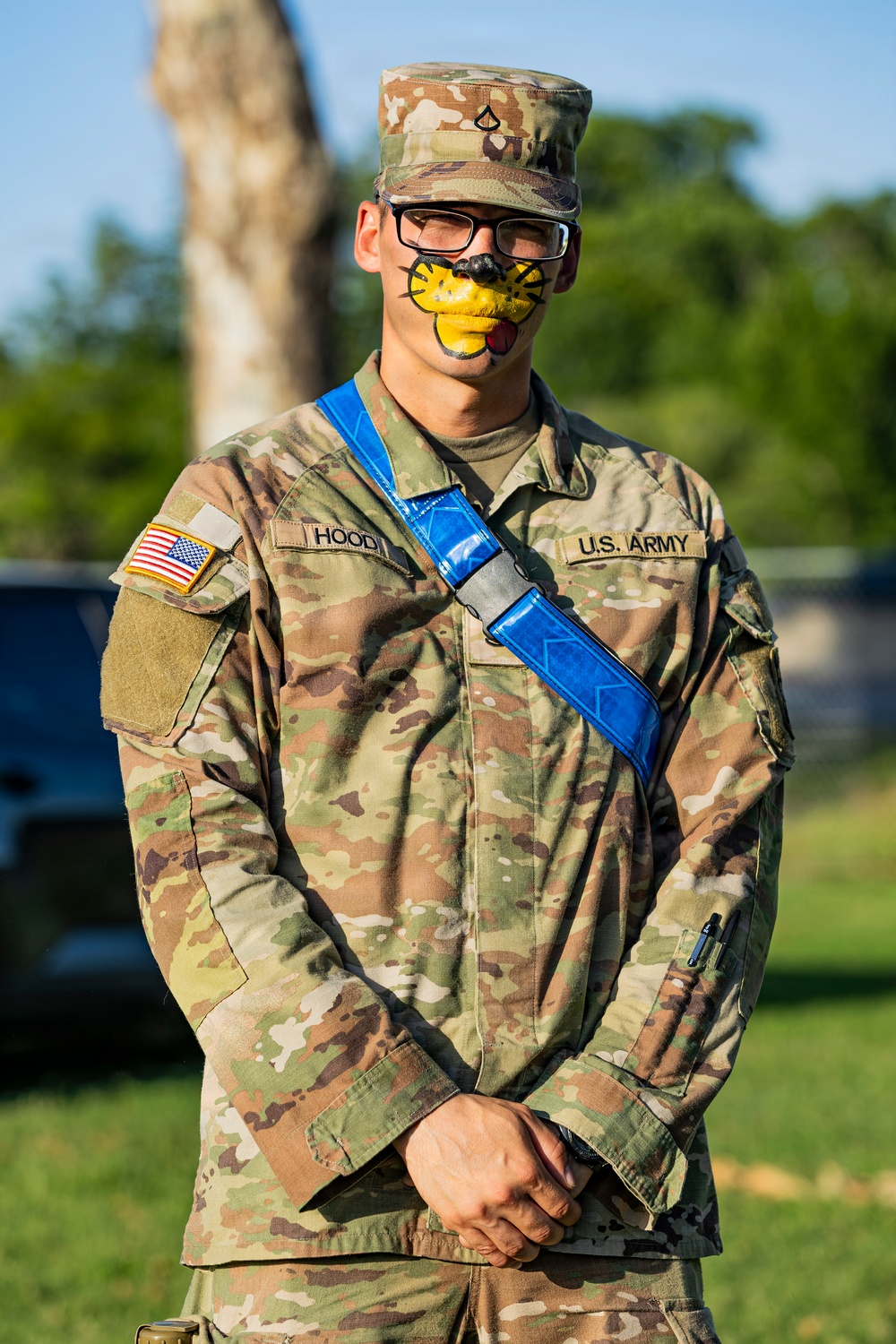 A U.S. Army soldier named Hood stands at attention in MultiCam (OCP) uniform with a blue diagonal sash/strap across the chest. The soldier has novelty face paint depicting a yellow duck or bird beak/mouth on the lower half of their face, suggesting a unit morale event, hazing tradition, or spirit activity. The soldier wears glasses and a patrol cap with a Private First Class rank insignia.