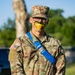 A U.S. Army soldier named Hood stands at attention in MultiCam (OCP) uniform with a blue diagonal sash/strap across the chest. The soldier has novelty face paint depicting a yellow duck or bird beak/mouth on the lower half of their face, suggesting a unit morale event, hazing tradition, or spirit activity. The soldier wears glasses and a patrol cap with a Private First Class rank insignia.