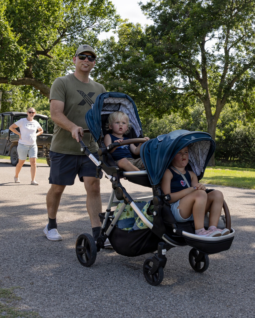 A man wearing a green t-shirt, baseball cap, and sunglasses pushes a navy blue double stroller containing two young toddlers on a paved path outdoors. A woman in a white shirt and shorts stands in the background near a utility vehicle. The setting appears to be a park or outdoor recreational area with large trees on a sunny day.