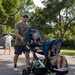 A man wearing a green t-shirt, baseball cap, and sunglasses pushes a navy blue double stroller containing two young toddlers on a paved path outdoors. A woman in a white shirt and shorts stands in the background near a utility vehicle. The setting appears to be a park or outdoor recreational area with large trees on a sunny day.