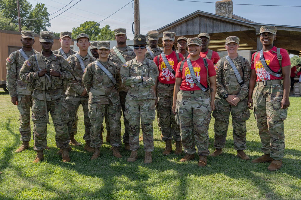 A group of approximately 14 military personnel posing together outdoors on a grassy area near a wooden structure. The group includes soldiers in full OCP (Operational Camouflage Pattern) uniforms and some individuals wearing red 'Freedom' event t-shirts with military pants. One individual in the center wears a distinctive drill sergeant campaign hat.