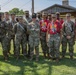 A group of approximately 14 military personnel posing together outdoors on a grassy area near a wooden structure. The group includes soldiers in full OCP (Operational Camouflage Pattern) uniforms and some individuals wearing red 'Freedom' event t-shirts with military pants. One individual in the center wears a distinctive drill sergeant campaign hat.