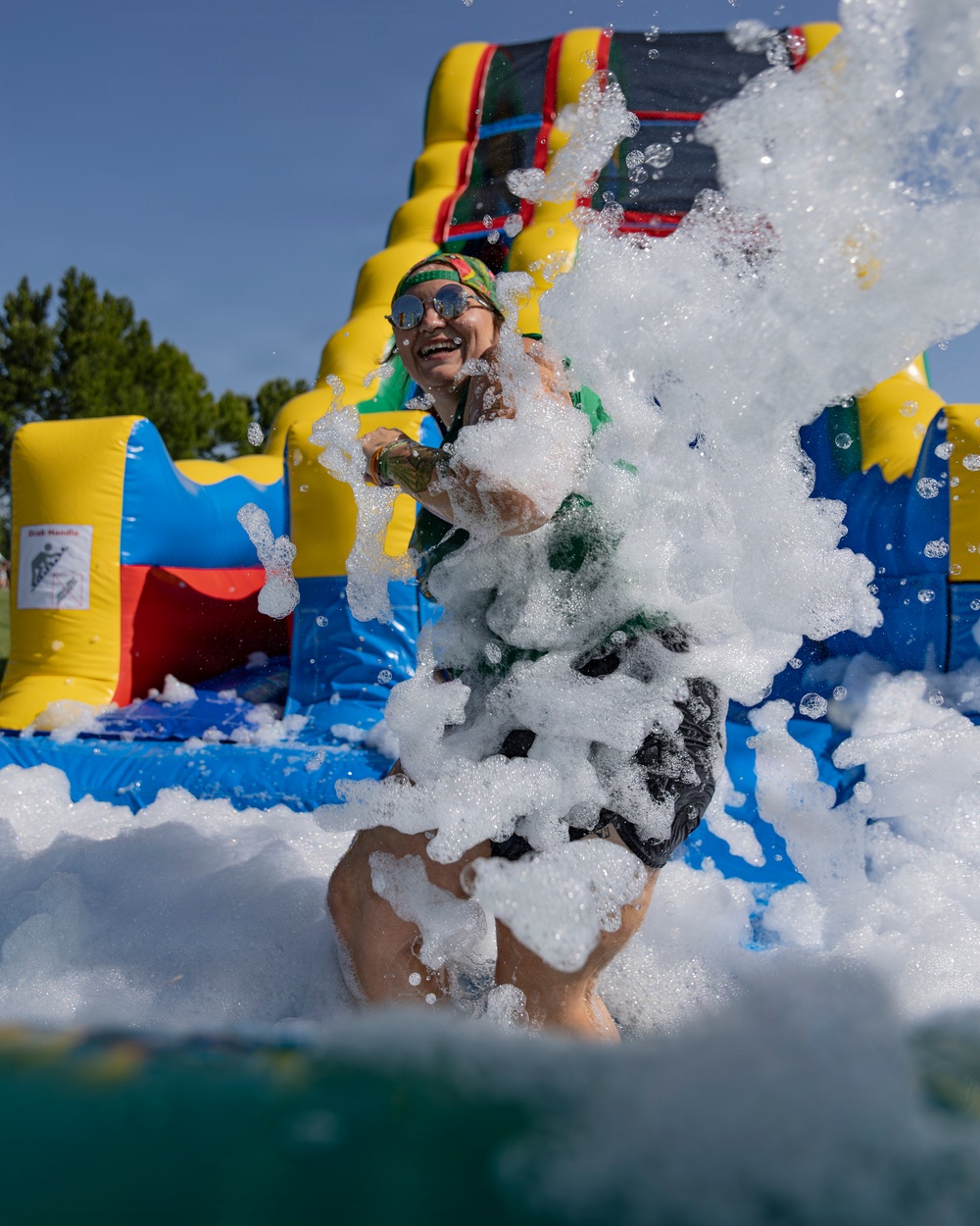 A person is playing in a foam pit at an outdoor inflatable water slide attraction on a sunny day. They are laughing and throwing foam into the air, surrounded by large white soap bubbles. The colorful inflatable structure in the background features yellow, blue, red, and green panels with a slide.