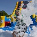 A person is playing in a foam pit at an outdoor inflatable water slide attraction on a sunny day. They are laughing and throwing foam into the air, surrounded by large white soap bubbles. The colorful inflatable structure in the background features yellow, blue, red, and green panels with a slide.