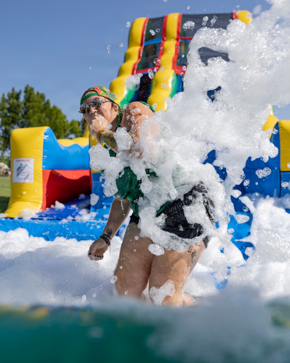 A person is captured mid-action sliding into a foam pit at an outdoor inflatable attraction event on a sunny day. They are surrounded by a large splash of white foam bubbles, wearing a green outfit, sunglasses, and a colorful head covering. Multiple colorful inflatable structures are visible in the background.