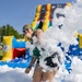 A person is captured mid-action sliding into a foam pit at an outdoor inflatable attraction event on a sunny day. They are surrounded by a large splash of white foam bubbles, wearing a green outfit, sunglasses, and a colorful head covering. Multiple colorful inflatable structures are visible in the background.