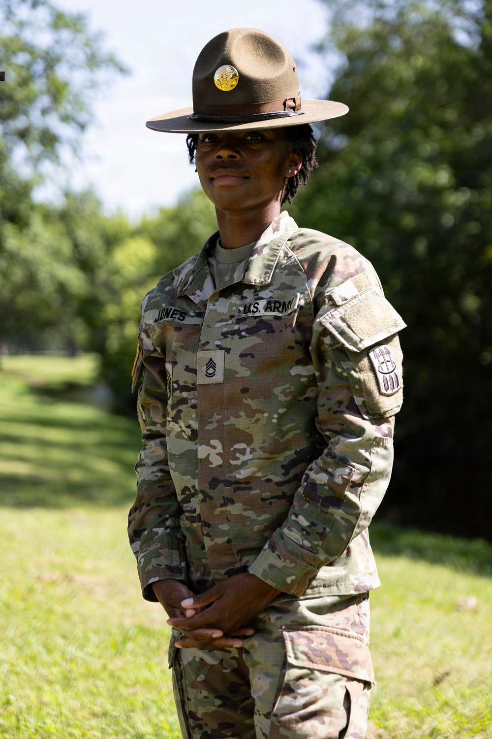 A U.S. Army Staff Sergeant named Jones stands at ease in an outdoor setting wearing the OCP (Operational Camouflage Pattern) uniform and a Drill Sergeant campaign hat. The soldier displays rank insignia consistent with Staff Sergeant (E-6) and wears unit patches on the left sleeve. The image appears to be an official or semi-official portrait taken in a grassy, tree-lined environment.