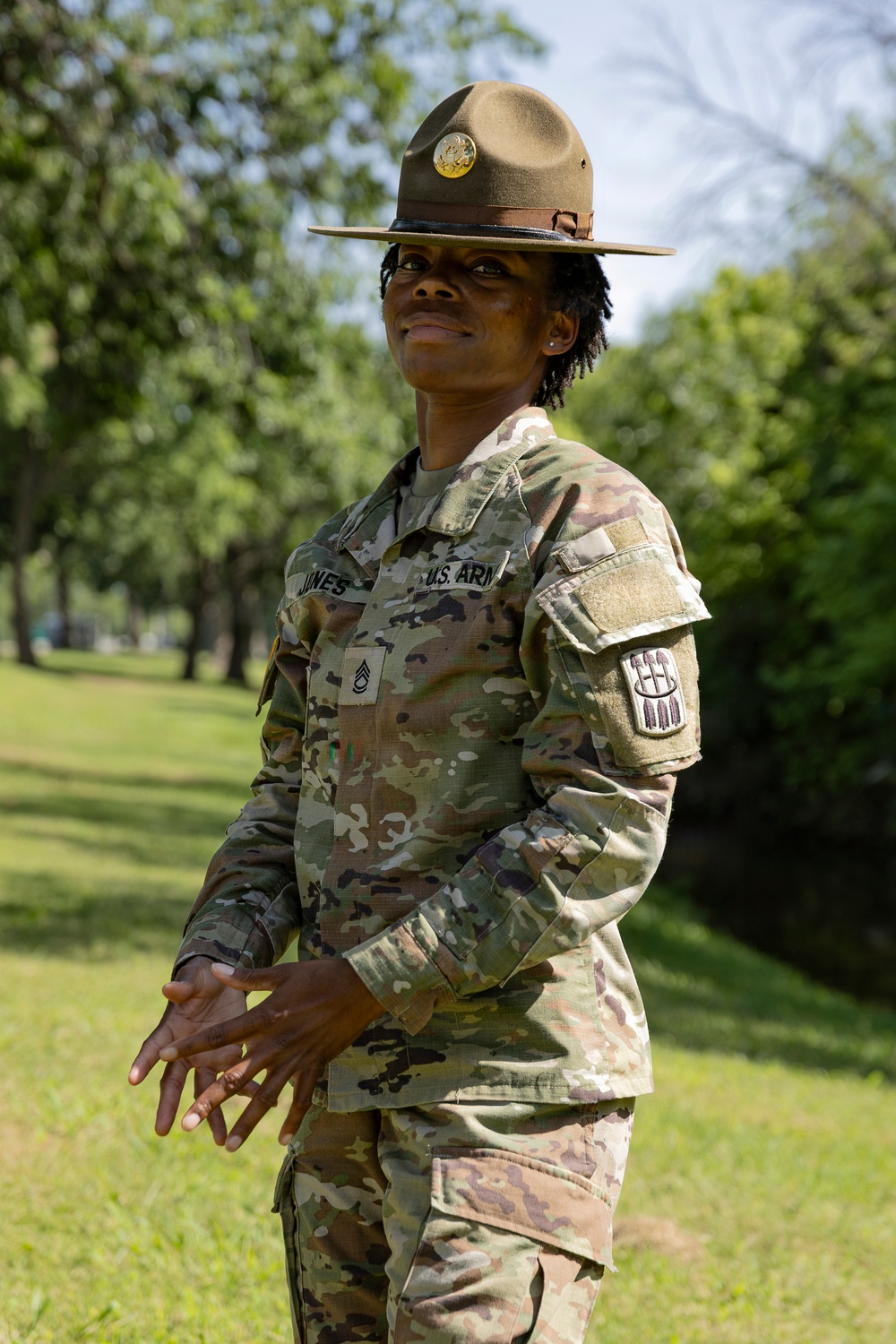 A U.S. Army Drill Sergeant stands outdoors in a grassy, tree-lined area wearing the distinctive campaign (Smokey Bear) hat and OCP (Operational Camouflage Pattern) uniform. The name tape reads 'JONES' and the rank insignia indicates Staff Sergeant. A unit patch is visible on the right sleeve.