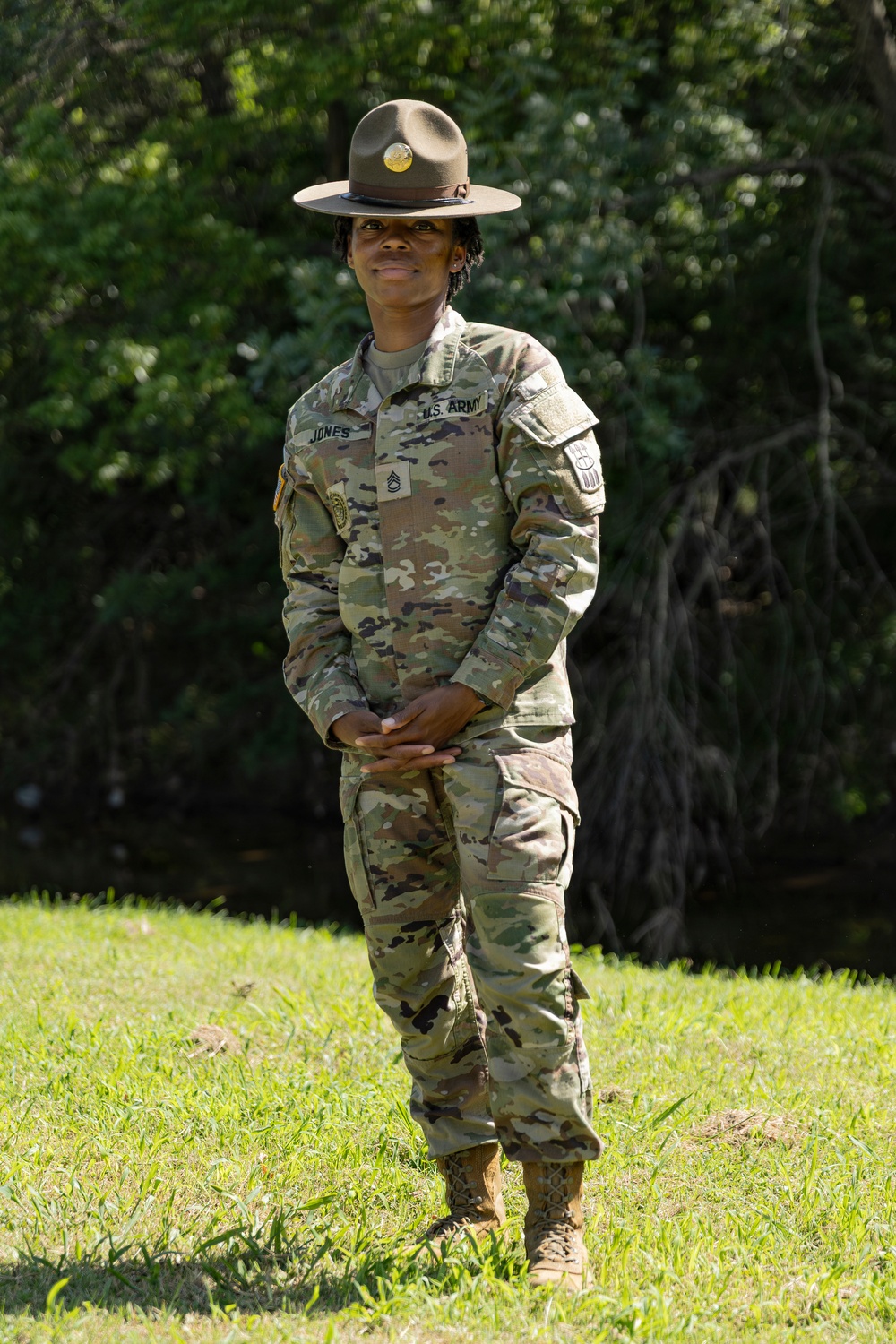 A U.S. Army Drill Sergeant named Jones stands at ease on a grassy field with wooded background. The soldier wears the distinctive Drill Sergeant campaign hat (Smokey Bear hat) along with the Army Combat Uniform (ACU) in OCP (Operational Camouflage Pattern). Rank insignia visible on the chest indicates the rank of Staff Sergeant (E-6).