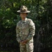 A U.S. Army Drill Sergeant named Jones stands at ease on a grassy field with wooded background. The soldier wears the distinctive Drill Sergeant campaign hat (Smokey Bear hat) along with the Army Combat Uniform (ACU) in OCP (Operational Camouflage Pattern). Rank insignia visible on the chest indicates the rank of Staff Sergeant (E-6).