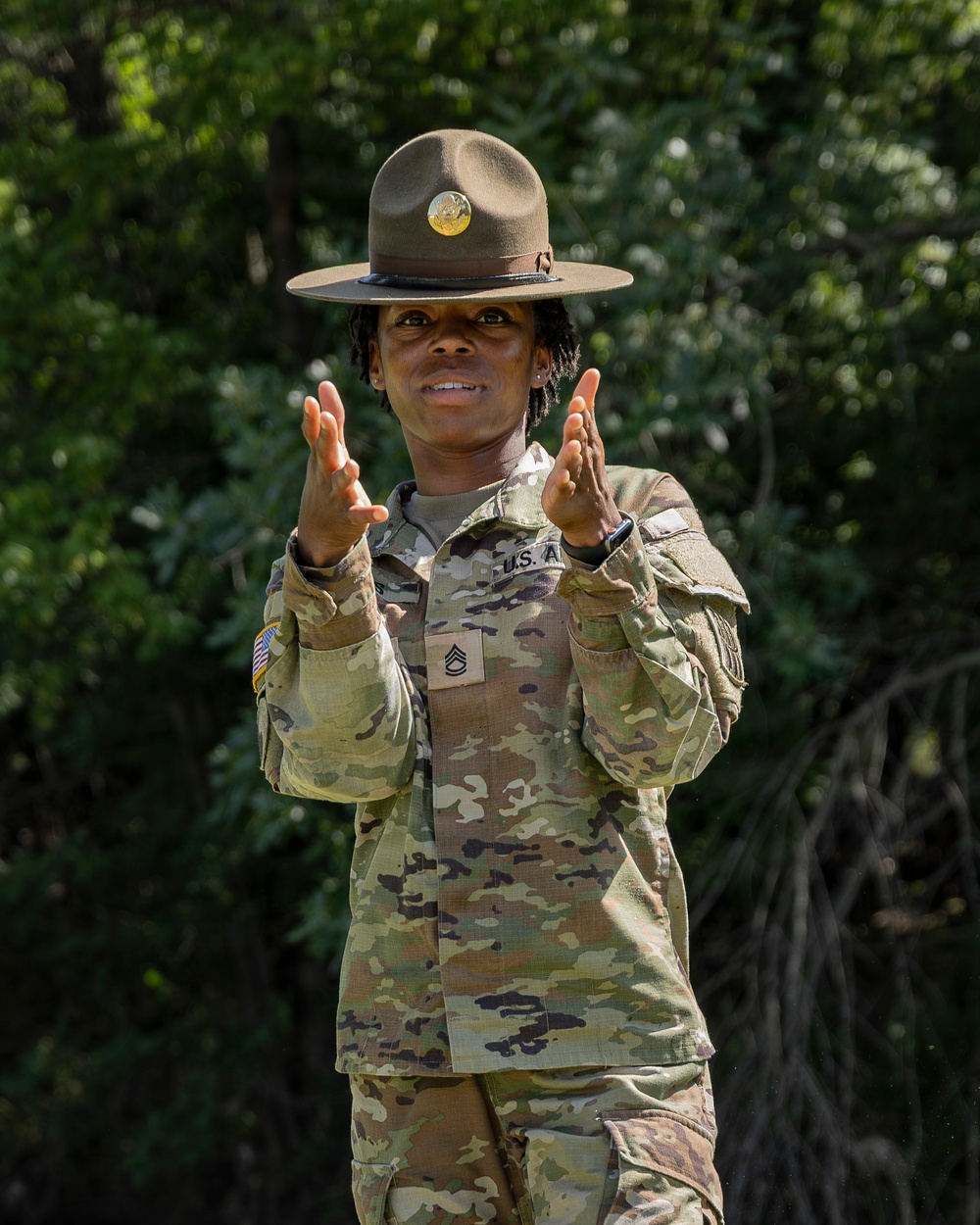 A U.S. Army Staff Sergeant wearing OCP (Operational Camouflage Pattern) uniform and a Drill Sergeant campaign hat is photographed outdoors against a green foliage background. The soldier has both hands raised in a gesturing position, suggesting active instruction or communication. The rank insignia and 'U.S. A[rmy]' tape are clearly visible on the uniform.