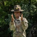 A U.S. Army Staff Sergeant wearing OCP (Operational Camouflage Pattern) uniform and a Drill Sergeant campaign hat is photographed outdoors against a green foliage background. The soldier has both hands raised in a gesturing position, suggesting active instruction or communication. The rank insignia and 'U.S. A[rmy]' tape are clearly visible on the uniform.