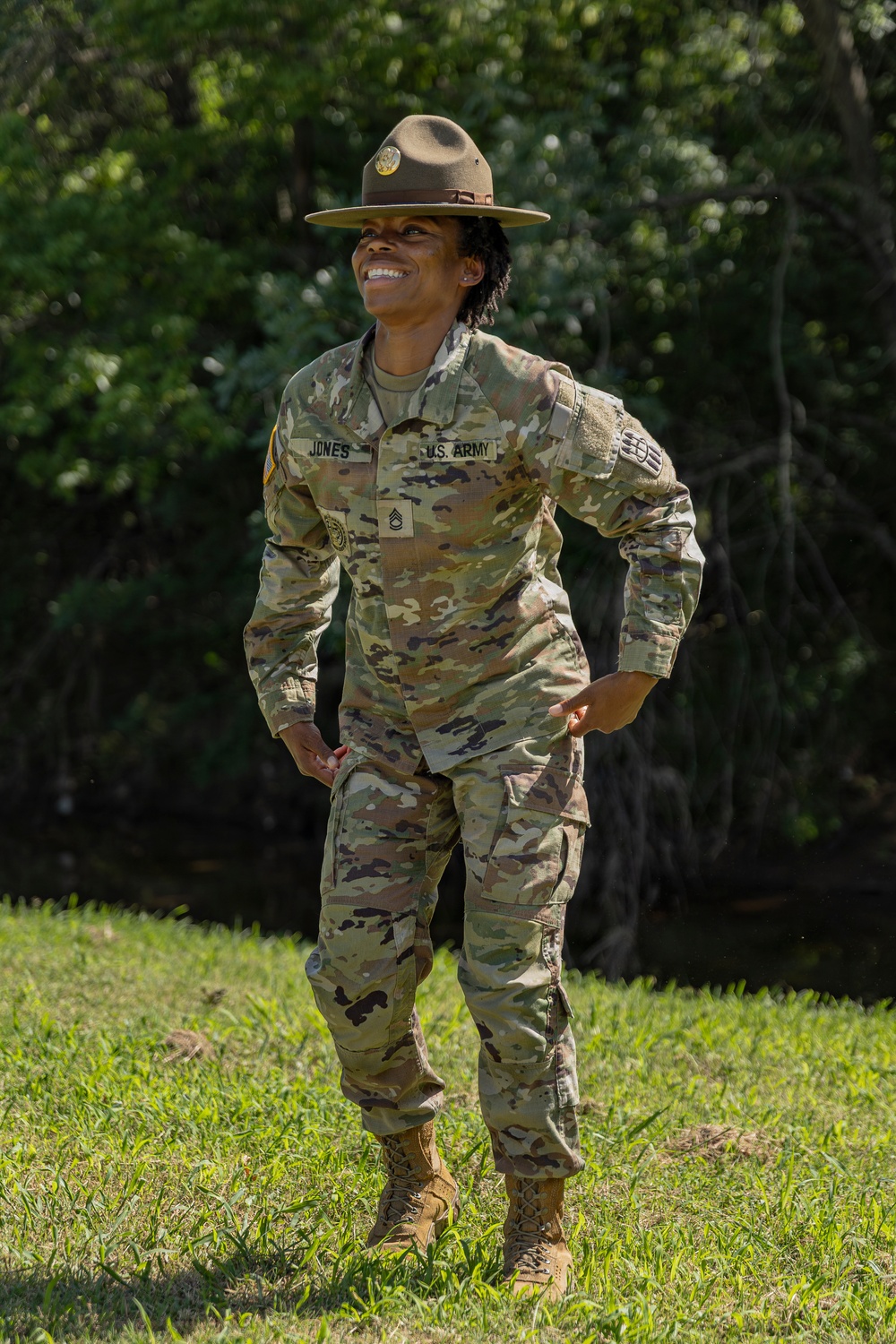 A U.S. Army soldier named Jones, wearing the OCP (Operational Camouflage Pattern) uniform, stands outdoors on grass with hands on hips and a broad smile. She is wearing a Drill Sergeant hat (campaign cover) and displays Staff Sergeant rank insignia on her chest. The setting appears to be a training or outdoor military environment with trees in the background.