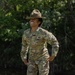 A U.S. Army soldier named Jones, wearing the OCP (Operational Camouflage Pattern) uniform, stands outdoors on grass with hands on hips and a broad smile. She is wearing a Drill Sergeant hat (campaign cover) and displays Staff Sergeant rank insignia on her chest. The setting appears to be a training or outdoor military environment with trees in the background.