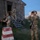 Deep Strike 10th Special Forces Group Three soldiers in multicam camouflage uniforms are conducting what appears to be a military training exercise at dusk near a weathered wooden structure. The personnel are equipped with rifles, night vision devices, and blue identification markers on their arms and helmets, indicating a training or exercise scenario. One soldier is using binoculars or a night vision monocular, one is holding a weapon in a ready position, and a third is ascending stairs into the building.