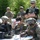 Deep Strike 10th Special Forces Group A group of military personnel in camouflage uniforms and tactical gear are gathered around a folding table conducting drone operations training or preparation. Three individuals in the foreground are actively working with a small quadcopter drone and associated equipment cases, while one operator wears FPV (first-person view) goggles. Three additional soldiers stand in the background observing the activity.