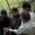 Deep Strike 10th Special Forces Group Four individuals are gathered in a wooded outdoor setting, appearing to review information on a handheld device. One person is wearing military multicam camouflage uniform, another wears civilian tactical clothing, and one individual's face has been digitally pixelated/redacted. The group appears to be engaged in a briefing or information-sharing activity in a forest environment.