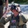 Deep Strike 10th Special Forces Group A U.S. military personnel wearing full tactical gear is adjusting a Skyzone FPV (First Person View) goggle headset, likely used for drone operation. The soldier is equipped with a tactical helmet mounted with camera/sensor equipment, body armor with an American flag patch, and communication devices. The setting appears to be an outdoor training or operational environment on a gravel road with trees in the background.