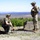 Deep Strike 10th Special Forces Group Two military personnel in tactical gear are operating a small drone on an elevated outdoor position with a wide valley and forested landscape visible in the background. One individual is crouching and handling a small quadcopter drone on the ground, while the other stands holding what appears to be a controller or device. The setting appears to be a training exercise or reconnaissance operation at a hilltop or elevated platform.
