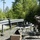 Deep Strike 10th Special Forces Group Two individuals, one in military tactical gear and one in civilian clothing, are operating drone and electronic equipment at an outdoor field station near a road guardrail. A folding table holds laptops, a drone, and various electronic components, while a separate tripod-mounted device is positioned to the left. The setting appears to be a wooded area during a military or joint exercise involving unmanned aerial systems.