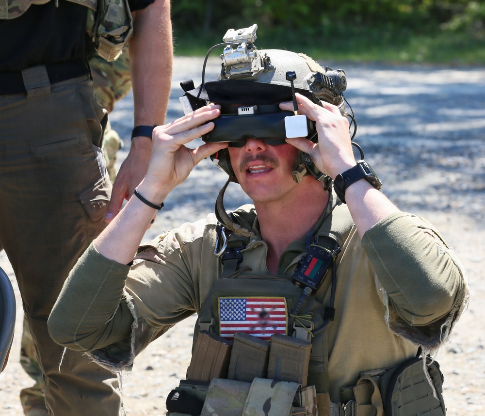 A U.S. military service member wearing tactical gear and a combat helmet is adjusting or fitting a heads-up display (HUD) or augmented reality visor device over their eyes. The soldier is equipped with an American flag patch on their plate carrier vest, along with various tactical accessories. The setting appears to be an outdoor training or demonstration environment on a gravel surface.