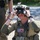 Deep Strike 10th Special Forces Group A U.S. military service member wearing tactical gear and a combat helmet is adjusting or fitting a heads-up display (HUD) or augmented reality visor device over their eyes. The soldier is equipped with an American flag patch on their plate carrier vest, along with various tactical accessories. The setting appears to be an outdoor training or demonstration environment on a gravel surface.