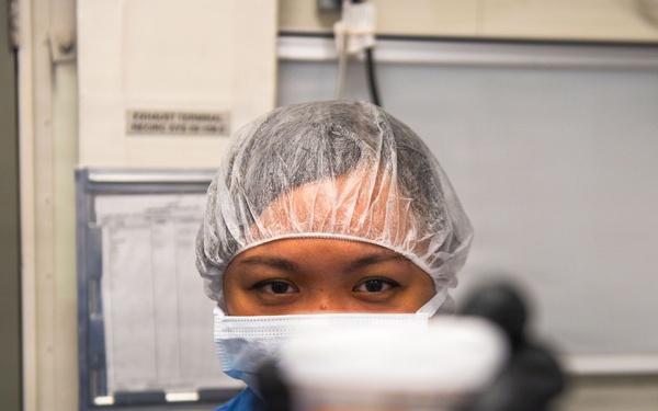 A healthcare or laboratory worker wearing blue scrubs, a hair net, face mask, and black nitrile gloves holds a specimen container toward the camera. The container appears to hold a red/crimson liquid, likely a biological specimen such as blood. The setting appears to be a clinical or laboratory environment.