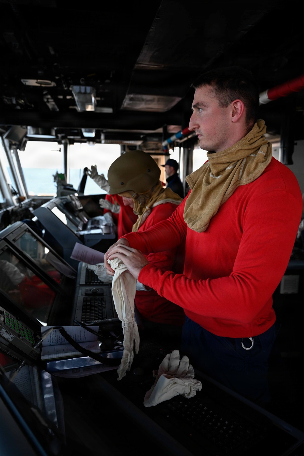 The image shows personnel on what appears to be a military or naval vessel's bridge/pilothouse. Two individuals in the foreground are wearing red long-sleeve shirts with tan neck gaiters, standing at navigation consoles. A third person is visible in the background near the bridge windows overlooking open water.