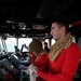 The image shows personnel on what appears to be a military or naval vessel's bridge/pilothouse. Two individuals in the foreground are wearing red long-sleeve shirts with tan neck gaiters, standing at navigation consoles. A third person is visible in the background near the bridge windows overlooking open water.